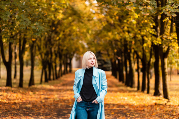Mode jeune femme en manteau bleu élégant avec pull noir et jeans promenades dans le parc avec des feuilles d'automne jaunes - Photo, image