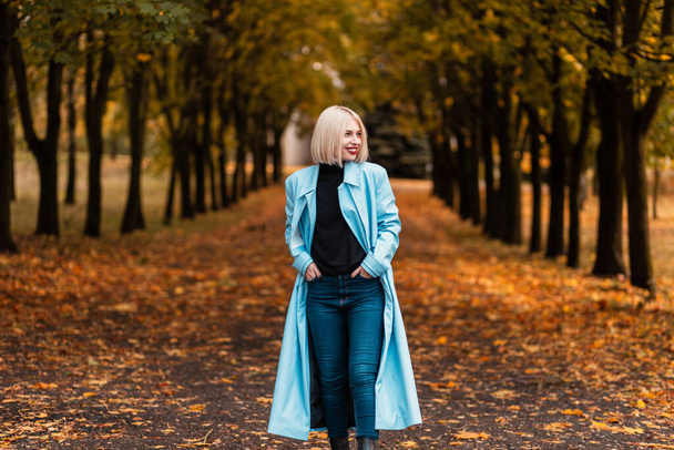Happy beautiful woman model with smile in fashion blue coat with jeans walks in autumn park with yellow fall leaves - Zdjęcie, obraz