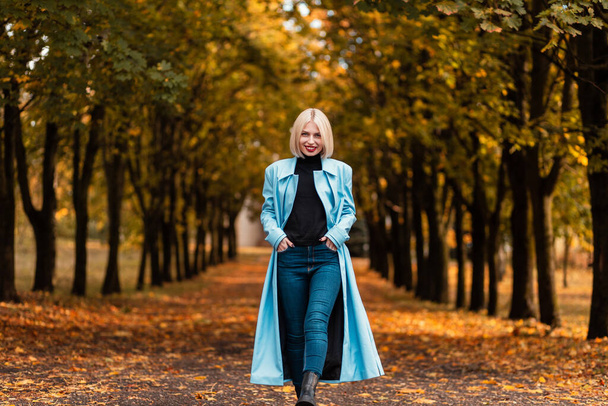 Šťastná podnikatelka v módních šatech s kabátem, svetrem a džínami procházky v podzimním parku s podzimními listy - Fotografie, Obrázek