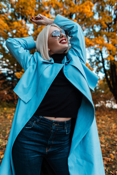 Feliz hermosa joven con gafas de sol en un abrigo azul de moda con un suéter negro y jeans en el bosque sobre un fondo de follaje amarillo de otoño - Foto, Imagen