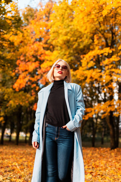 ブルーのおしゃれなコートにセーターとジーンズを身にまとい、ヴィンテージのサングラスをかけた美しい若い女性が秋の森の中を歩く。 - 写真・画像