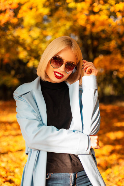 Jesienny portret pięknej młodej blondynki w okularach przeciwsłonecznych w jasnoniebieskim płaszczu z czarnym swetrem spacery w jesiennym parku ze złotymi liśćmi jesieni - Zdjęcie, obraz