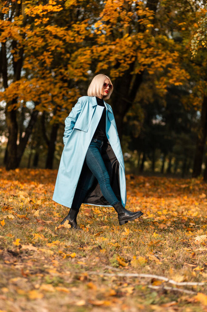 Elegante modelo de mujer rubia elegante con abrigo azul de moda camina en un increíble parque de otoño con follaje dorado brillante - Foto, imagen