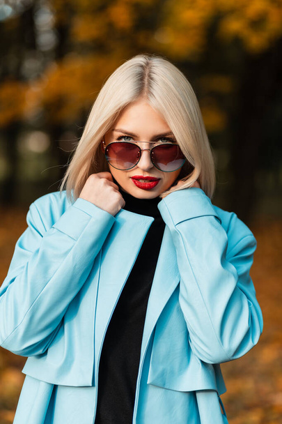 Autumn portrait of a beautiful blonde woman with red lips in fashion clothes with sunglasses on the background of an fall park with bright colored foliage - Fotó, kép