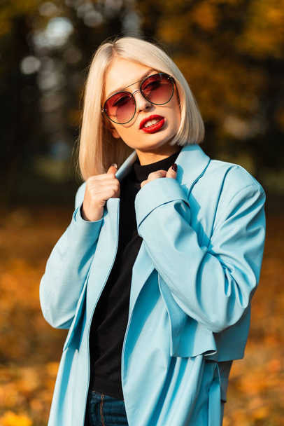 Stylová krásná mladá dívka s červenými rty a slunečními brýlemi v módním modrém kabátě se svetrem procházky v parku se zlatým podzimním listím - Fotografie, Obrázek