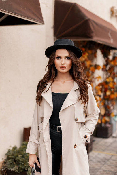 Gyönyörű fiatal nő göndör hajjal, divatos kalapban, klasszikus szürke kabátban sétál a városban egy őszi napon. - Fotó, kép
