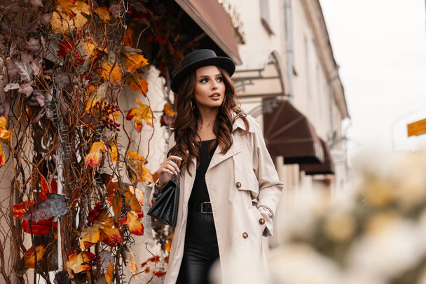 Portrait d'une jolie femme à la mode avec un chapeau élégant dans un manteau gris à la mode avec un sac à main se promène dans la ville avec un feuillage d'automne jaune - Photo, image