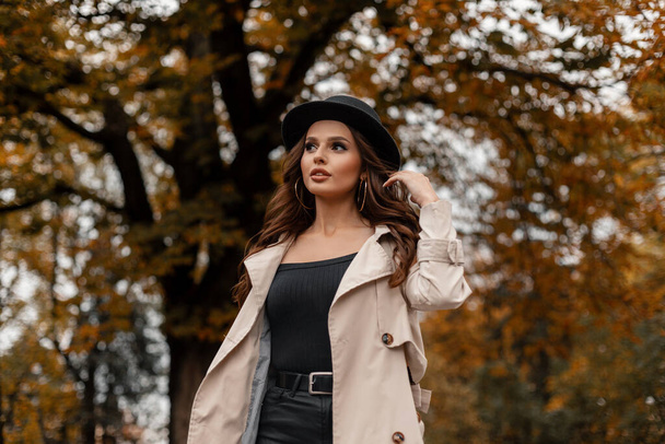 Модная красивая юная стройная девушка в модной шляпе в стильном сером пальто ходит по улице на фоне деревьев с желтой осенней листвы - Фото, изображение