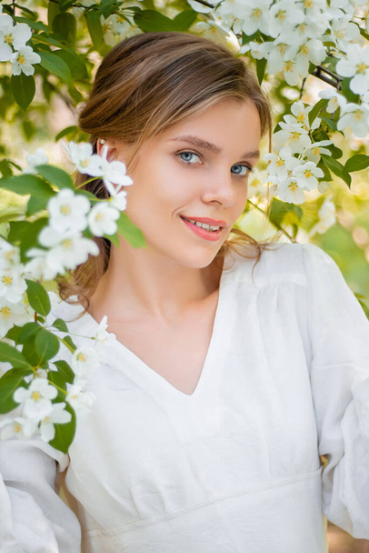 Portrait of a young woman in a white dress against the backdrop of blooming apple trees. The girl poses against the background of flowers in the spring park. - Foto, imagen
