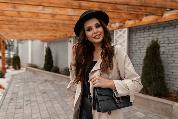 Glückliches junges, lockiges, brünettes Mädchen mit einem Lächeln in modischer Kleidung mit Vintage-Hut und Lederhandtasche geht auf die Straße. Eleganter weiblicher Stil und Schönheit im Herbst - Foto, Bild