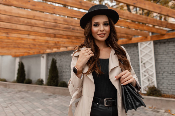 Stylish beautiful sexy young woman with a sweet smile in a fashion hat and gray classic coat with a leather black bag walks in the city - Fotoğraf, Görsel