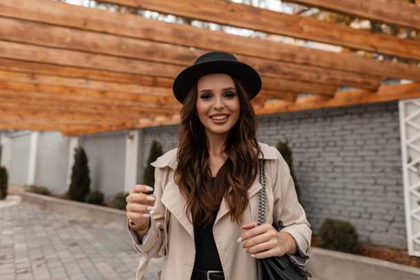 ritratto di una bella giovane donna bruna riccia con un sorriso carino in un cappello alla moda con un elegante cappotto vintage e una borsa di pelle cammina per strada. Stile femminile elegante, moda e bellezza - Foto, immagini