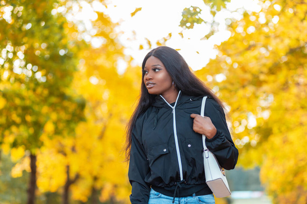 秋のカナダ公園を歩くハンドバッグ付きのファッショナブルなジャケットを着た美しい若い黒人アフリカ人女性明るい黄色の葉 - 写真・画像