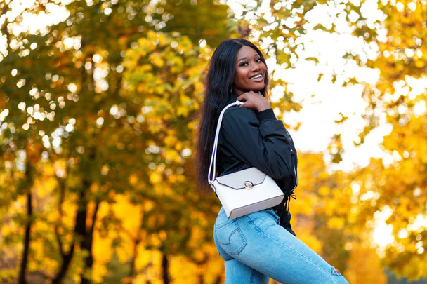 Feliz hermosa chica negra joven con una sonrisa en una chaqueta de moda y vaqueros azules con estilo con un bolso de cuero blanco en un parque canadiense con hojas de arce amarillo brillante otoño - Foto, Imagen