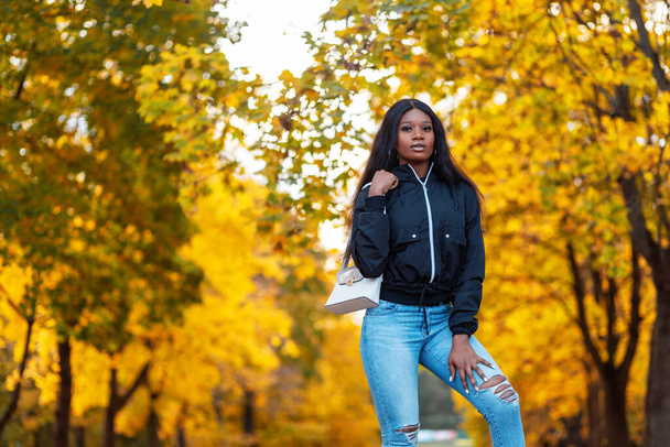 Fashion black american woman model in stylish casual jacket with blue jeans and bag walks in amazing autumn park with yellow fall leaves - Photo, image
