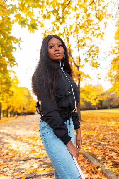 Elegante chica negra hermosa en ropa de moda con una chaqueta, jeans y un bolso blanco en la naturaleza con follaje de otoño amarillo brillante - Foto, Imagen