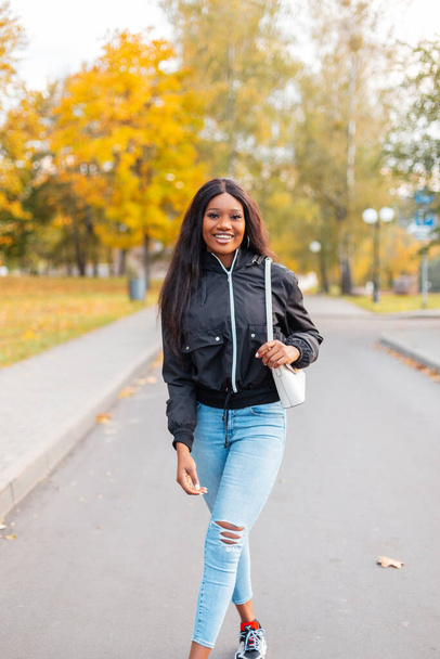 Chica negra bastante sonriente con ropa casual de moda con una chaqueta de moda, jeans y un elegante bolso está caminando en un parque de otoño con follaje de otoño amarillo brillante - Foto, Imagen