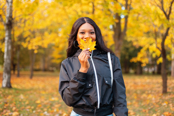 Feliz joven sonriente chica negra en chaqueta casual de moda cubre su cara con hoja de otoño amarillo en el parque con follaje dorado - Foto, Imagen