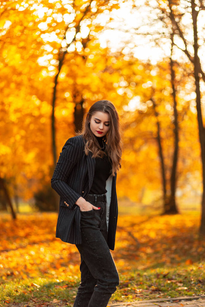jong mooi meisje in een zwart stijlvol pak met een modieuze blazer op de natuur met heldere herfst bladeren bij zonsondergang - Foto, afbeelding