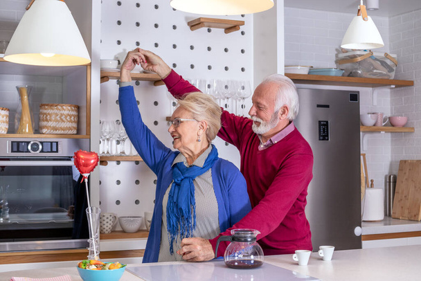 Glückliche Senioren tanzen Hand in Hand in der Küche. Ehemann gratuliert seiner Frau zum Valentinstag oder zum Hochzeitstag. - Foto, Bild
