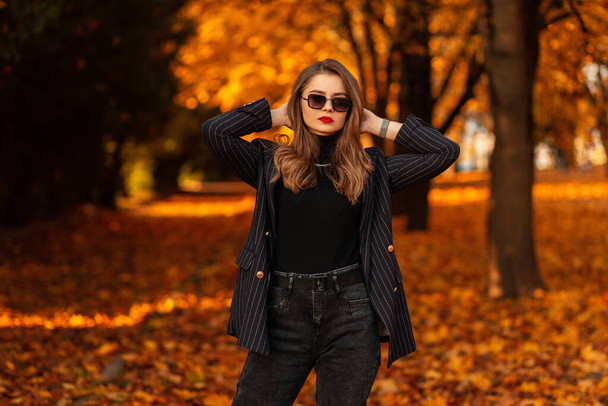 Stílusos gyönyörű fiatal nő divatos őszi ruhák egy kabát, pulóver és napszemüveg pózol egy parkban fényes őszi színű lombozat. Női üzleti stílus és szépség - Fotó, kép