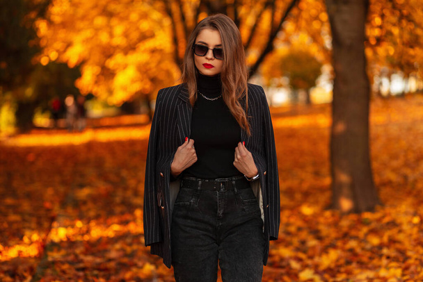 Elegante chica bonita con gafas de sol vintage en un traje elegante negro de moda con una chaqueta camina en la naturaleza con colorido follaje naranja de otoño - Foto, Imagen