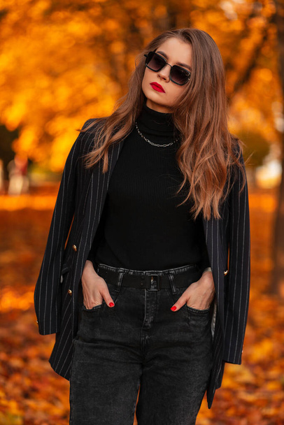 beautiful young business woman with sunglasses in a stylish suit with a fashionable blazer walks in an autumn park with an orange leaves - Foto, Imagem