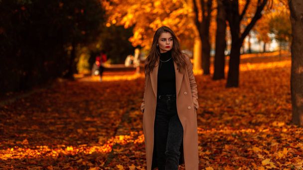 秋の公園を歩く黒いセーターを着たスタイリッシュなベージュのコートに赤い唇を持つファッショナブルな女性日没時にオレンジの葉 - 写真・画像