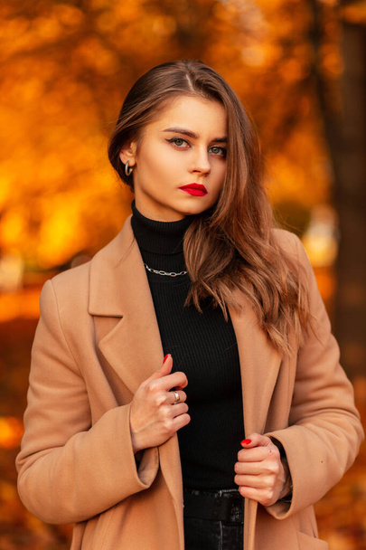 Porträt einer schönen jungen Frau mit roten Lippen in einem modischen beigen Mantel mit Pullover in einem Park mit farbigem gelben Herbstlaub. Weiblicher lässiger Stil und Schönheit - Foto, Bild