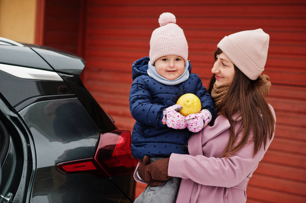 Young mother with baby girl on hands against car, hold lemon at winter. - Foto, imagen