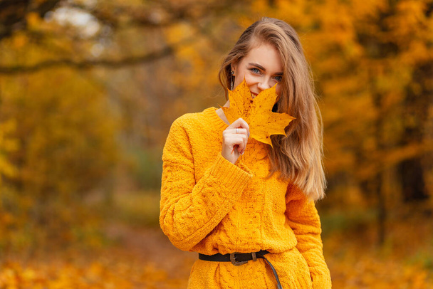 Beautiful young fashionable woman with a smile in a yellow vintage sweater covers her face with an autumn golden leaf on nature with orange fall foliage. Enjoy a happy moments - Photo, Image