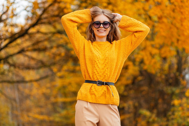 Joyeux beau contenté jeune fille avec un sourire dans des lunettes de soleil à la mode dans un pull jaune tricoté marche dans la campagne avec des feuilles d'automne orange - Photo, image