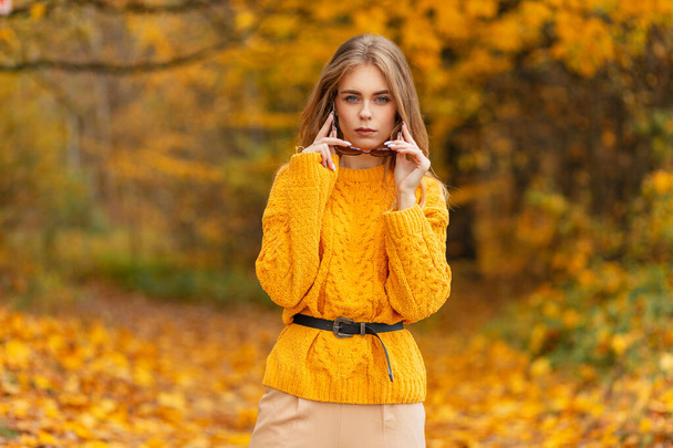 Moda hermosa mujer joven de moda en punto vintage suéter amarillo vestidos gafas de sol al aire libre sobre fondo de follaje de otoño dorado - Foto, Imagen