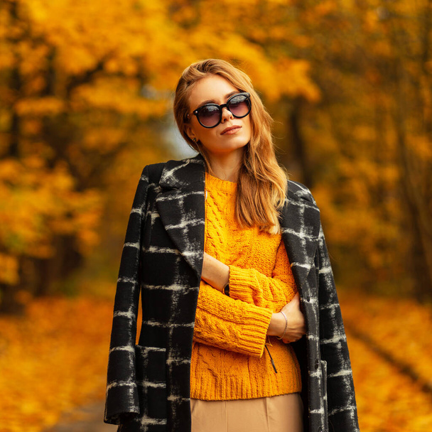 Schöne junge kaukasische Frau in einem gestrickten gelben Pullover und einem modischen Mantel mit stilvoller Sonnenbrille an einem strahlenden Herbsttag. Gelb bunte Herbst-Stil - Foto, Bild