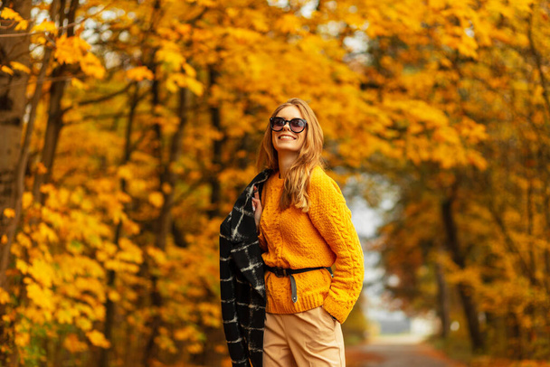 Pretty good woman with beautiful smile in fashion autumn clothes with knitted yellow vintage sweater and black coat walks outdoors with amazing fall leaves and trees - Photo, Image