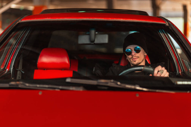 Hipster άνθρωπος με γυαλιά ηλίου κάθεται πίσω από το τιμόνι ενός κόκκινου παλιού αυτοκινήτου. Ταξίδι με αυτοκίνητο - Φωτογραφία, εικόνα