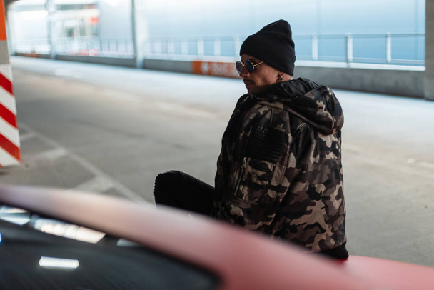 Стильный красивый молодой хипстер в солнечных очках и черной шляпе в модном зимнем военном пиджаке сидит на красном автомобиле в городе - Фото, изображение