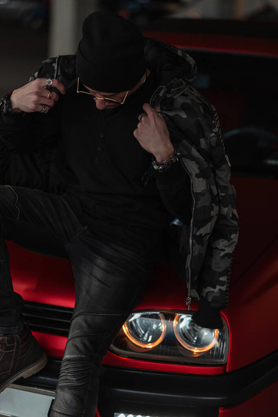 beau jeune modèle guy hipster avec un chapeau et des lunettes de soleil vintage dans une veste militaire noire avec un pull et jeans est assis sur une voiture dans la ville - Photo, image