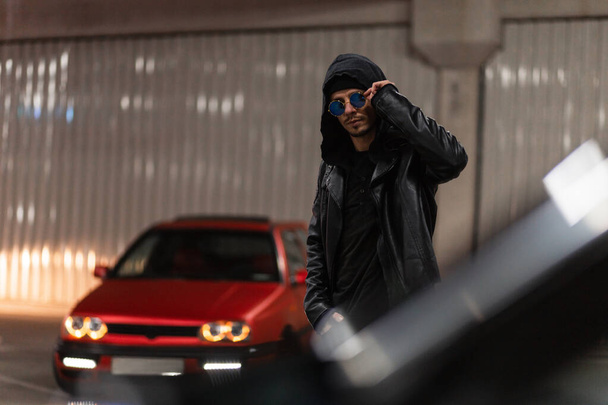 Fashionable young hipster guy with sunglasses in stylish clothes look with a jacket and a hoodie walks near the cars in the night parking - Photo, Image