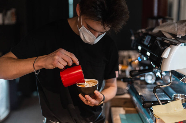 Barista suojaavassa lääketieteellisessä naamiossa mustassa t-paidassa tekee herkullista cappuccino-kahvia maidolla kahvilassa - Valokuva, kuva