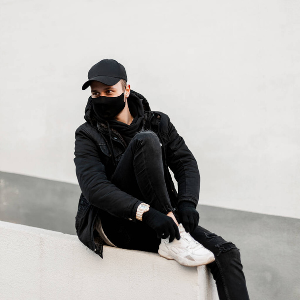 Μοντέρνος άνδρας με ιατρική μάσκα σε κομψά μαύρα ρούχα με σακάκι, καπέλο, κουκούλα και λευκά sneakers κάθεται κοντά σε ένα λευκό τοίχο στην πόλη - Φωτογραφία, εικόνα