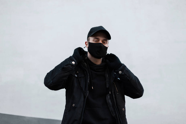 Cooler Hipster-Mann mit medizinischer Schutzmaske und schwarzer Mütze in modischer schwarzer Kleidung mit Jacke und stylischem Kapuzenpullover in der Nähe der weißen Mauer auf der Straße. Urban modern Pandemie männlichen Stil - Foto, Bild