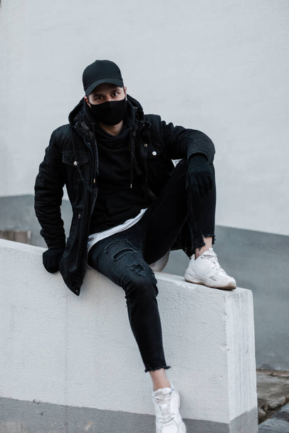 Tipo de moda en ropa elegante negro con gorra de diseño negro, chaqueta, sudadera con capucha, jeans y zapatillas blancas con una máscara protectora sentado fuera - Foto, imagen