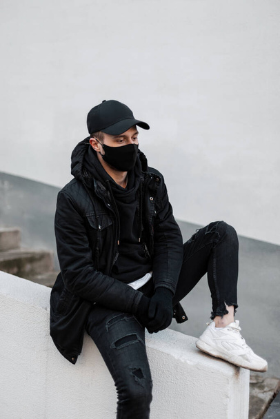 Modelo masculino bonito elegante com uma máscara protetora e boné em uma jaqueta na moda e um capuz com tênis se senta na rua perto de uma parede branca - Foto, Imagem