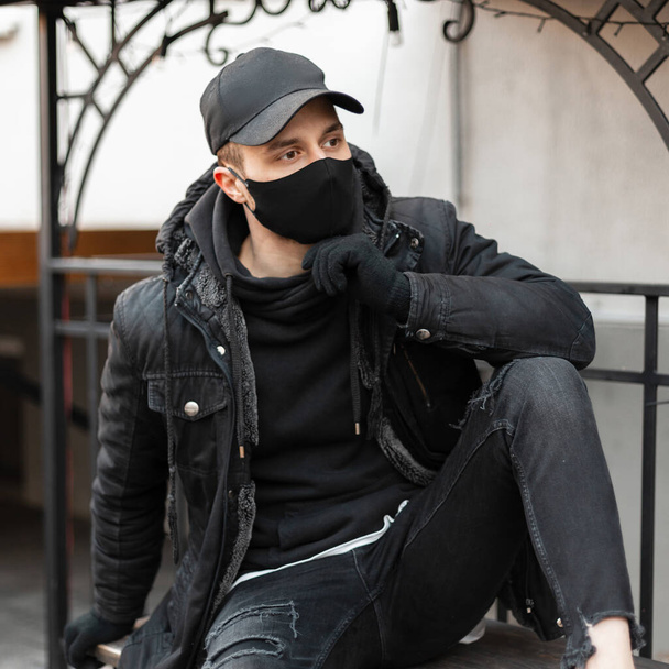 Fashionable handsome hipster guy with a black stylish cap and a protective medical mask in a black jacket and a hoodie sits in the city. Urban male casual modern style - Foto, Imagen