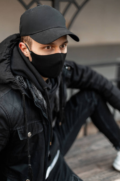 Fashionable handsome young man with protective black mask in fashion black jacket and hoodie with cap sits on the street - Photo, Image