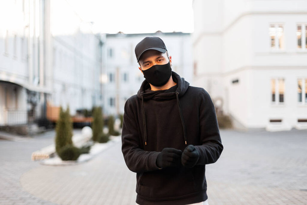 防護医療マスクとファッショナブルな黒いパーカーと街の手袋でスタイリッシュなキャップを持つハンサムな若いモデルの男。男性の近代的な都市型とパンデミック - 写真・画像