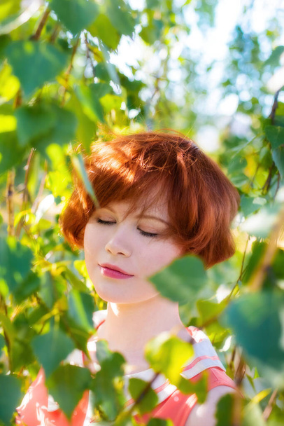 Portrait of a red-haired happy woman in park among the green foliage. - Фото, изображение