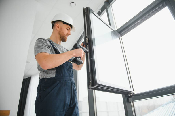 Workman in overalls installing or adjusting plastic windows in the living room at home - Фото, зображення