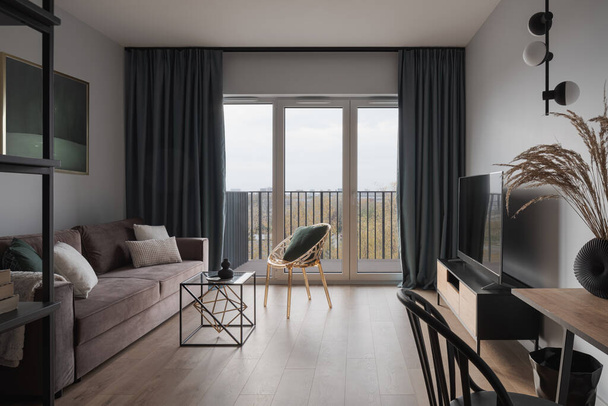 Проста і стильна вітальня з затишним диваном, сучасними меблями, телевізором і вікнами на балкон
 - Фото, зображення